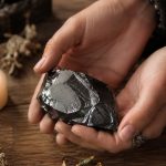 Hands holding a raw hematite stone beside a lit candle on a wooden table with dried herbs.