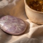 Close-up photograph of a pale lilac lepidolite palm stone on crumpled linen beside a ceramic cup of chamomile tea, lit by warm evening light with soft shadows and shallow depth of field