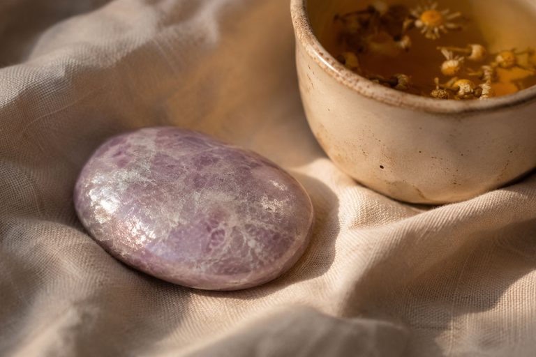 Close-up photograph of a pale lilac lepidolite palm stone on crumpled linen beside a ceramic cup of chamomile tea, lit by warm evening light with soft shadows and shallow depth of field