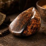 Polished bronzite stone on a dark table beside a candle, shield ornament, and dried herbs