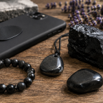 Shungite bracelet, pendant, and polished stones beside a smartphone on a wooden table with soft natural light and dried lavender