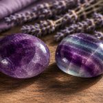 Two polished purple crystals, amethyst and fluorite, resting on a wooden table with dried lavender in the softly blurred background
