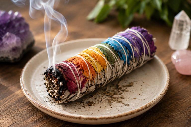Vibrant chakra-themed sage incense stick with smoke on a ceramic plate.