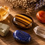 A tiger's eye palm stone at center surrounded by companion crystals — citrine, carnelian, pyrite, lapis lazuli, clear quartz — on a warm wooden surface